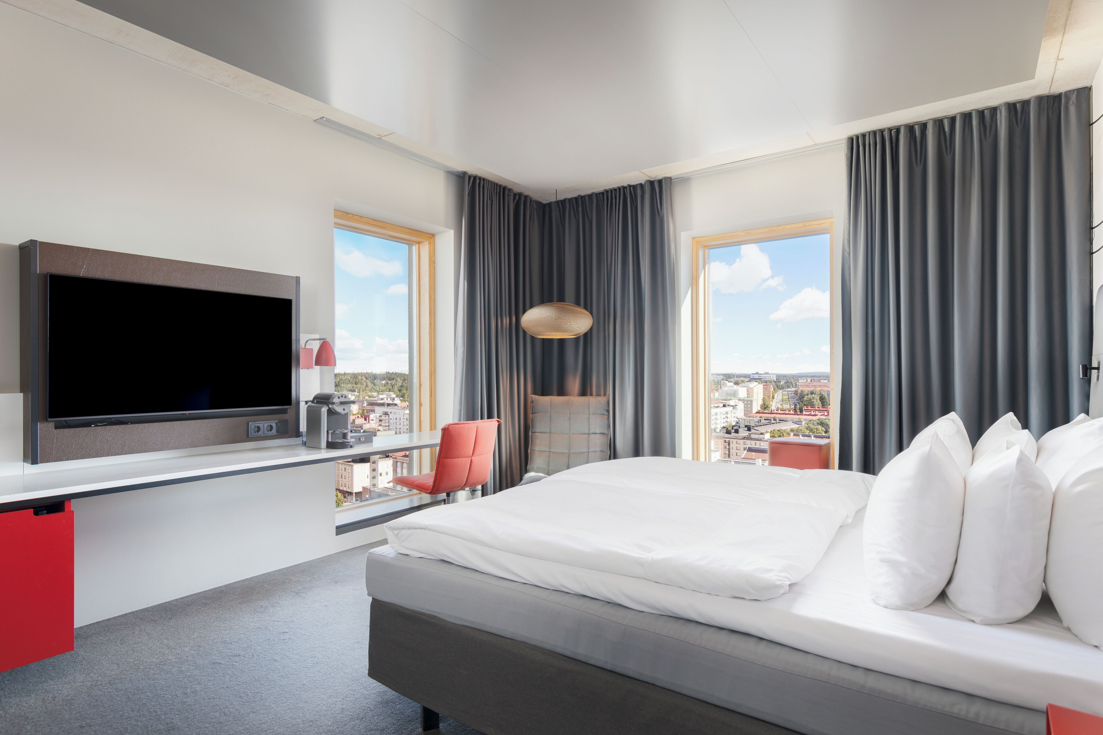 A modern hotel room with a large bed, a flat-screen TV, a desk with a red chair, and a city view through large windows.