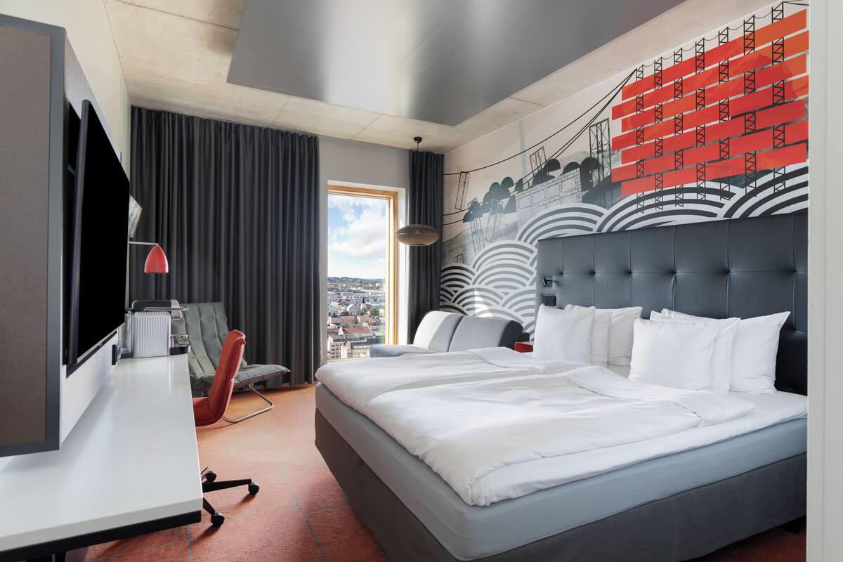 A modern hotel room with a large bed, a desk with a computer, a chair, and a view of the city.
