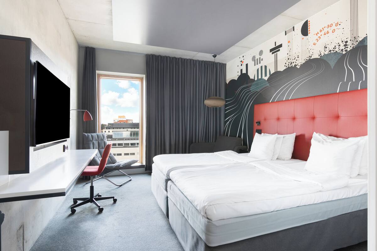 Ein Hotelzimmer mit Bett, Schreibtisch, Stuhl, Fernseher und einem Fenster mit Blick auf Gebäude.