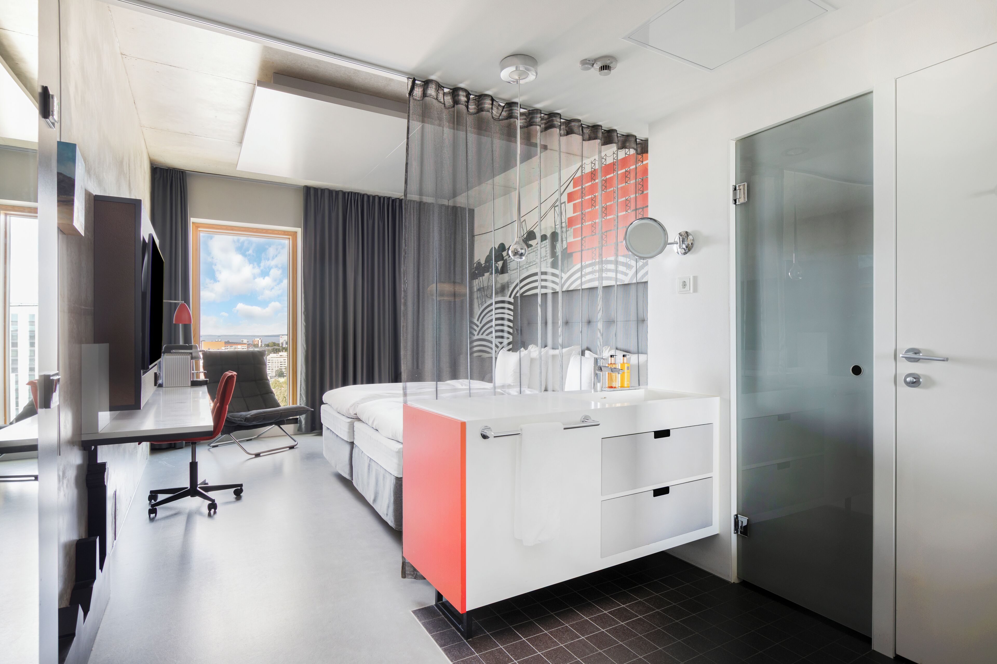 A modern hotel room with a large bed, a desk with a chair, a glass shower enclosure, and a view of the city.