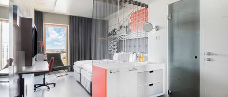 A modern hotel room with a large bed, a desk with a chair, a glass shower enclosure, and a view of the city.