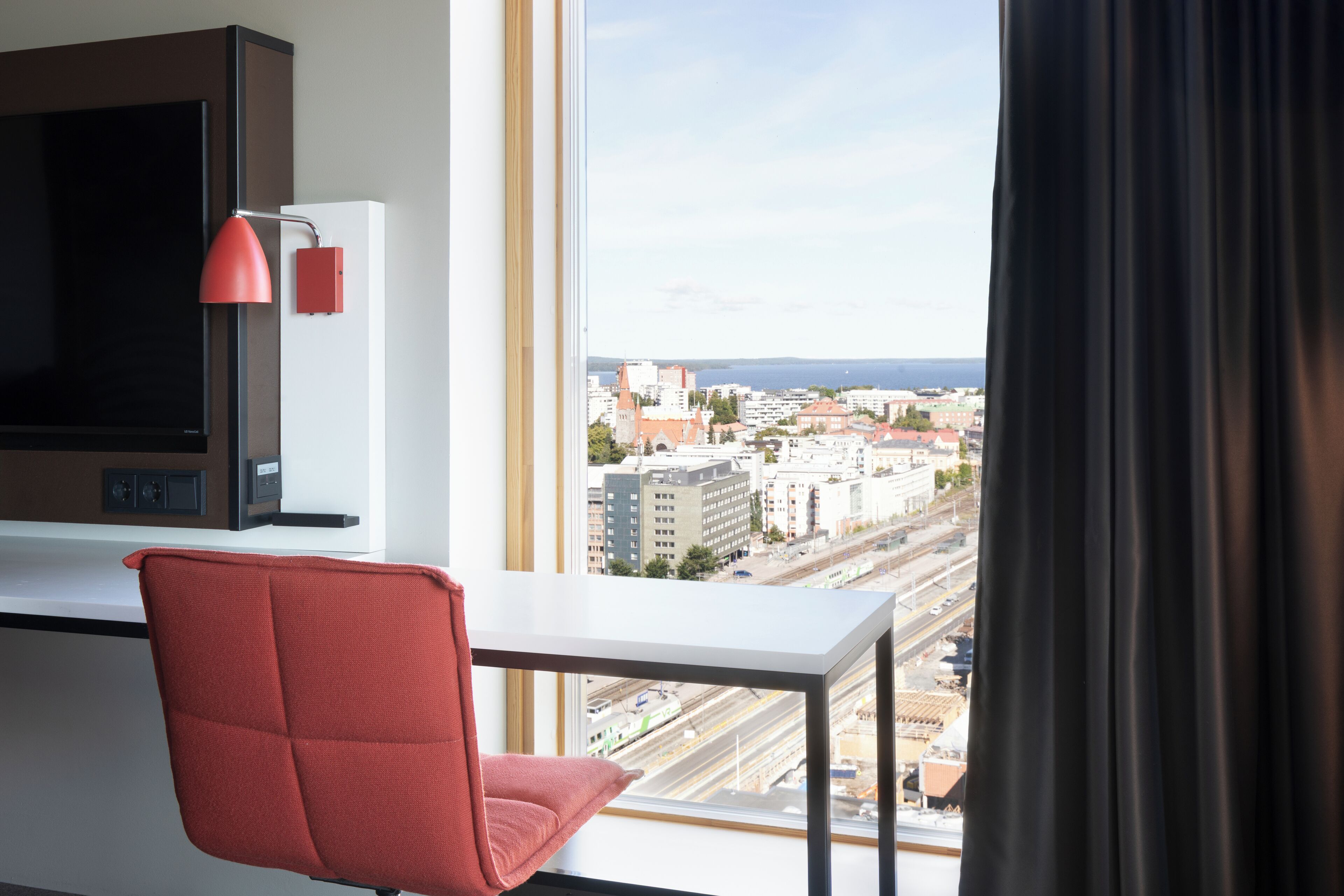 Une chambre d’hôtel avec une grande fenêtre, un fauteuil rouge, un téléviseur à écran plat et une lampe de bureau rouge.