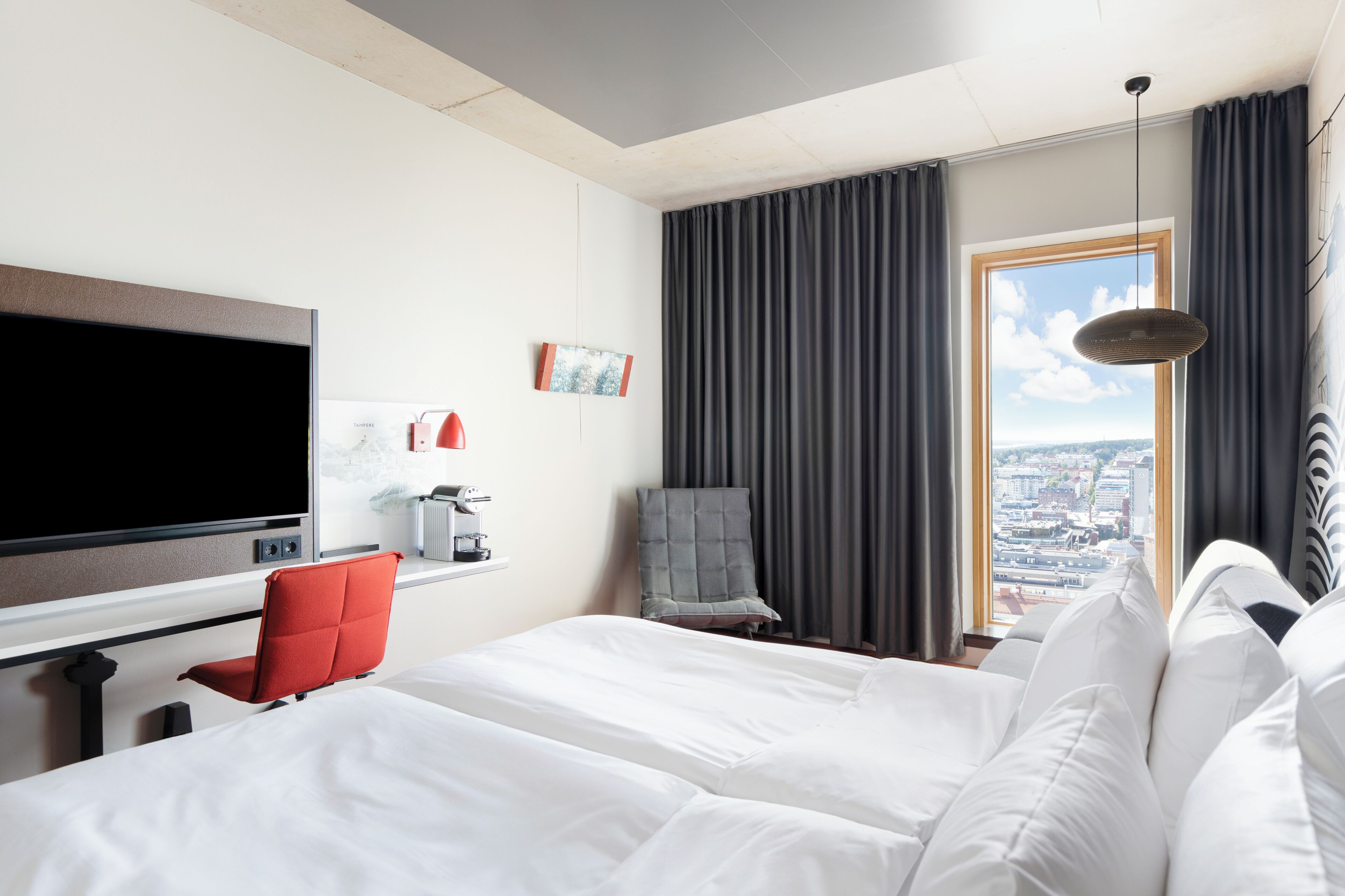Una camera d'albergo con un letto grande, una televisione, una macchina del caffè, una poltrona rossa e vista sulla città dalla finestra.
