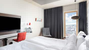A hotel room with a large bed, a television, a coffee machine, a red chair, and a view of the city through a window.