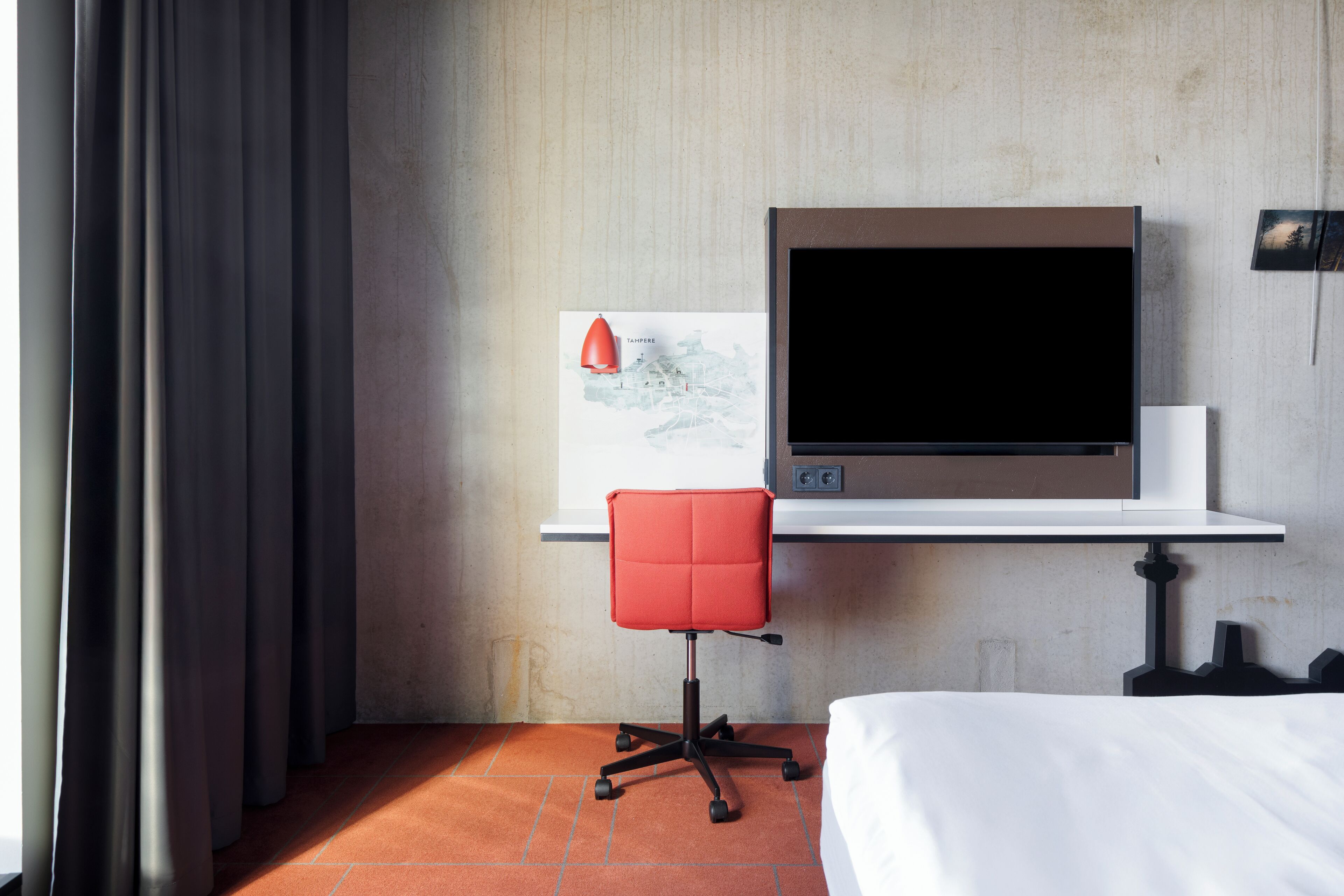 Ein Hotelzimmer mit einem Bett, einem Schreibtisch mit Computerbildschirm, einem roten Bürostuhl und einer Karte an der Wand.