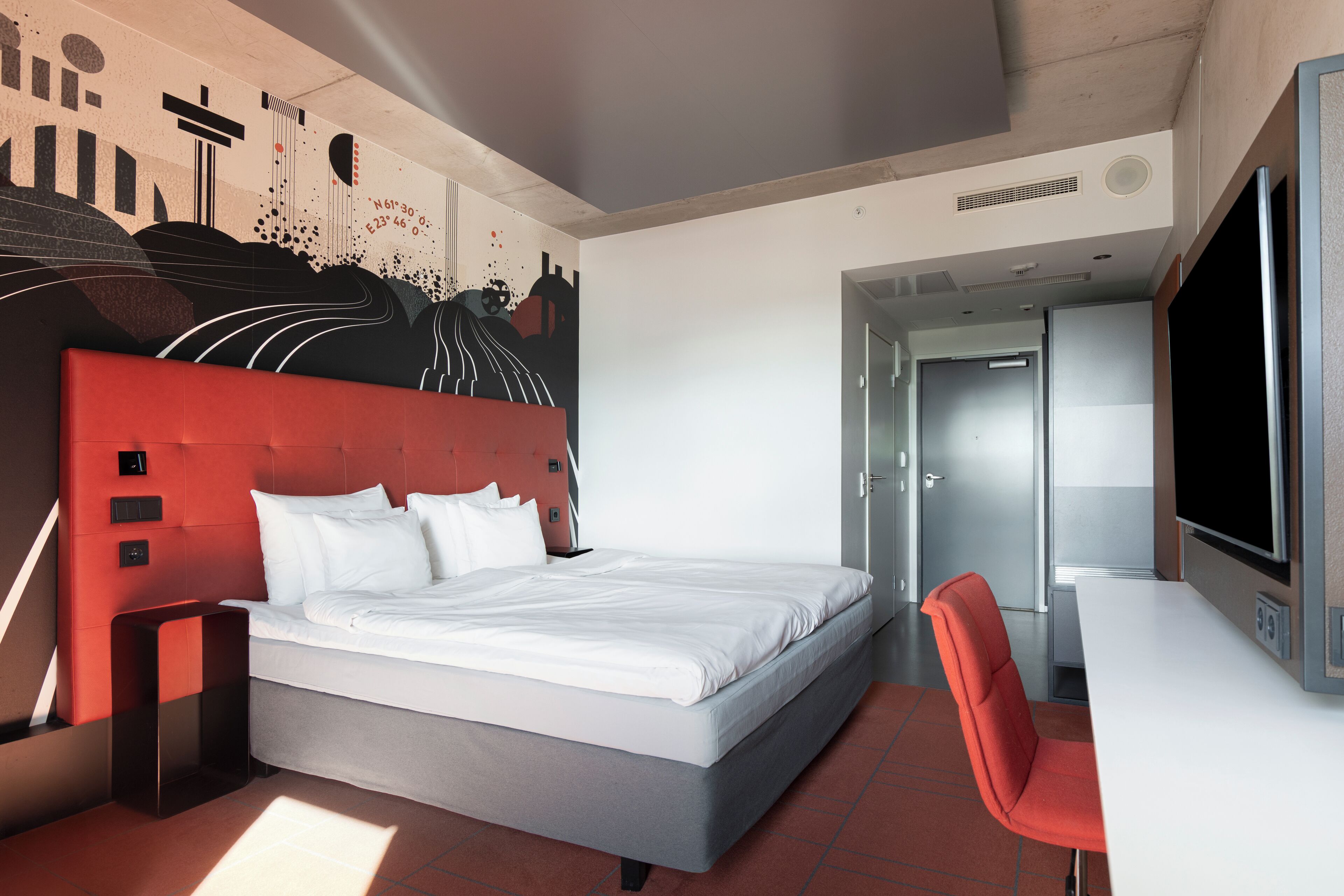 A modern hotel room with a large bed, a red accent wall, a desk, and a television.