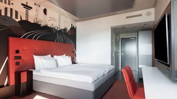 A modern hotel room with a large bed, a red accent wall, a desk, and a television.