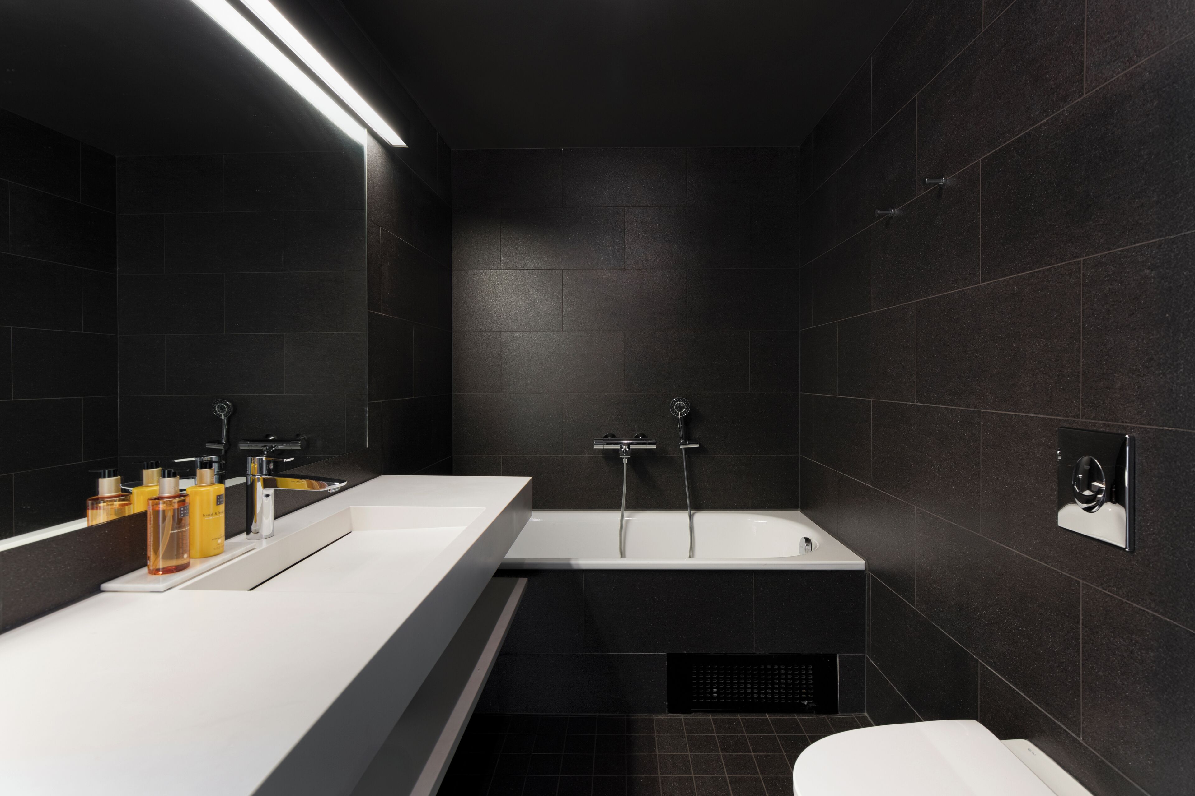 Een moderne badkamer met donkere tegels, een witte wastafel en een bad.