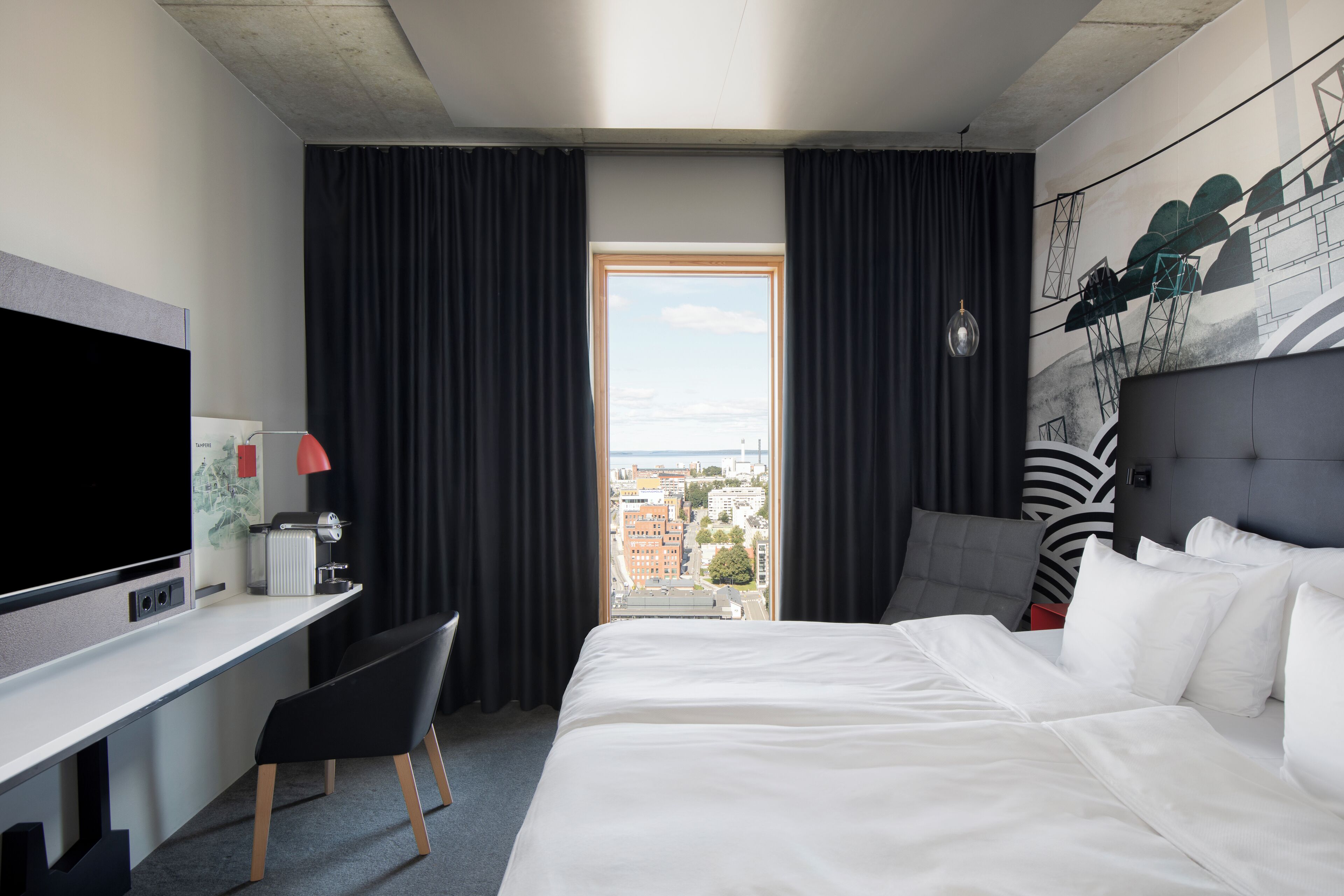 Una habitación de hotel con cama, escritorio con televisor, silla, ventana con cortinas y vistas a la ciudad.