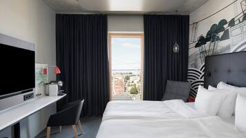 Una habitación de hotel con cama, escritorio con televisor, silla, ventana con cortinas y vistas a la ciudad.