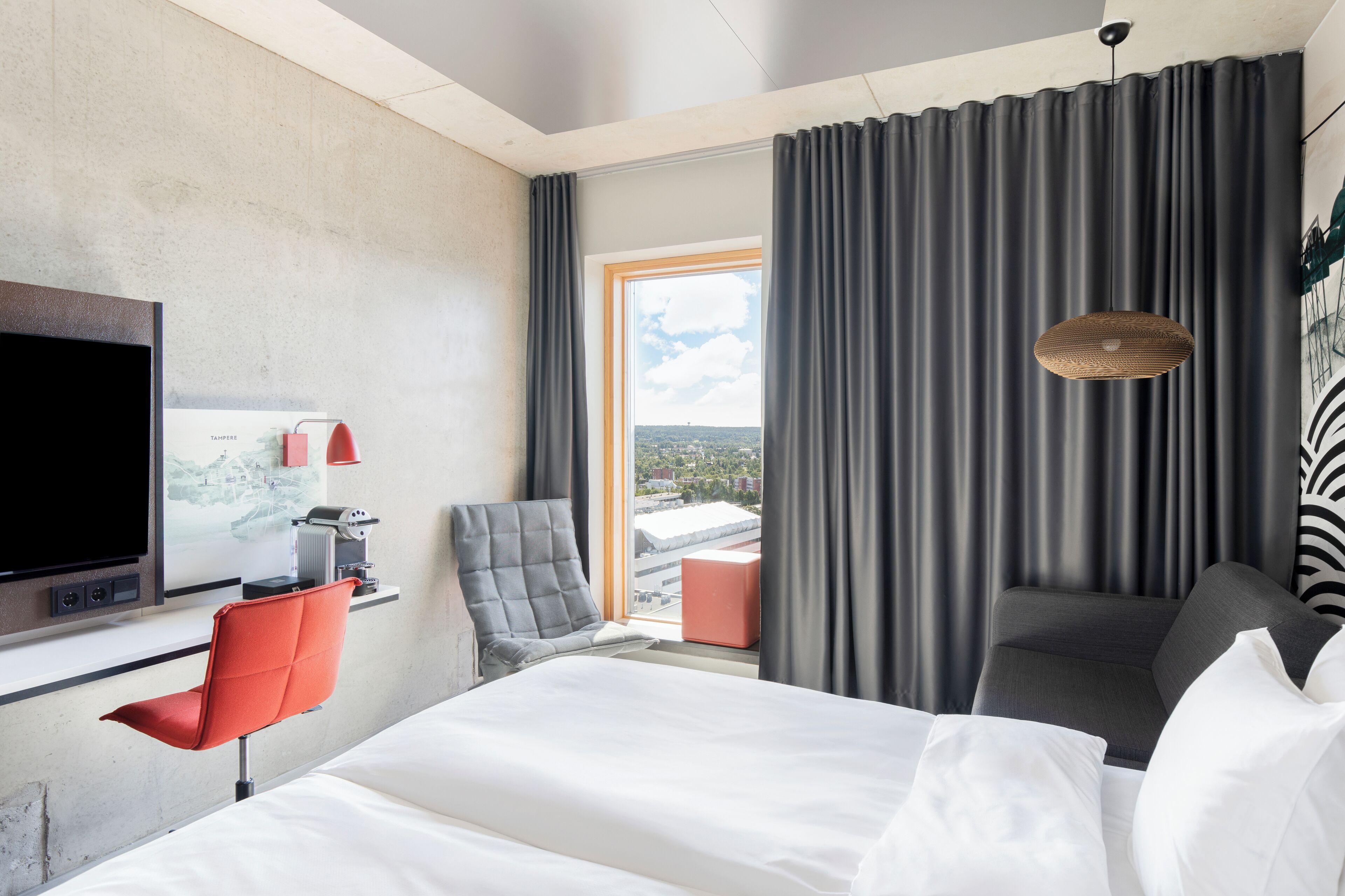Habitació d'hotel amb un llit, una televisió, una taula amb un ordinador, una cadira, una finestra amb cortines i un llum penjant.