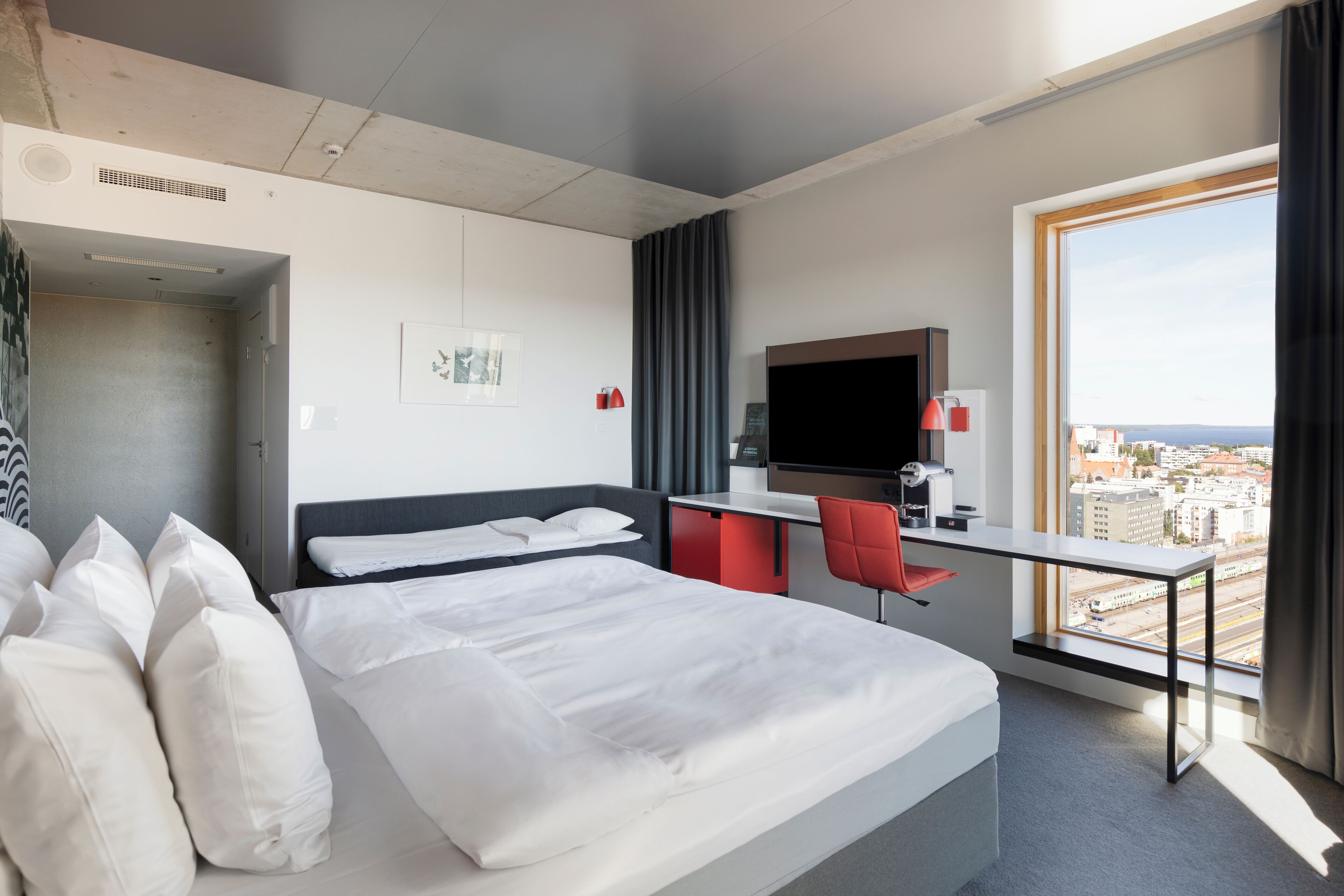 A hotel room with a bed, a desk, a chair, a TV, and a large window with a city view.