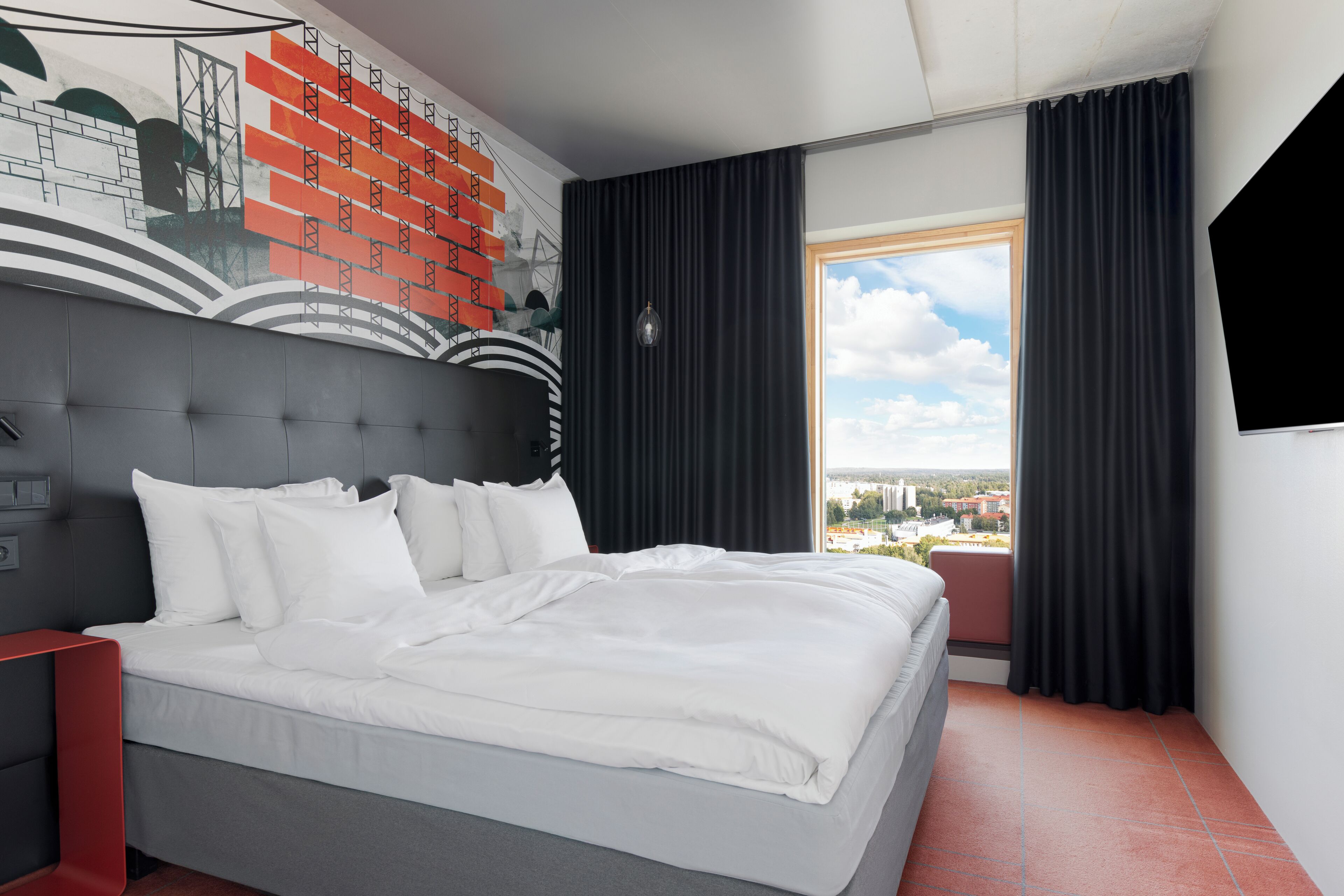 Una habitación de hotel moderna con una cama grande, una mesita de noche roja, un mural en la pared y vistas a la ciudad a través de la ventana.