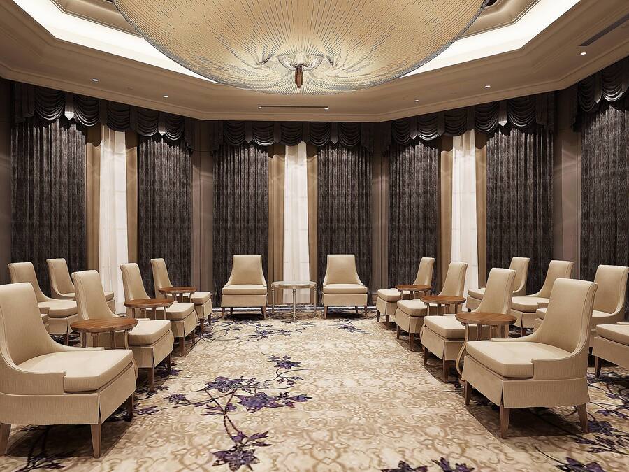 Un salón de conferencias con filas de sillas y mesas color beige, una alfombra estampada y un techo con una lámpara central.