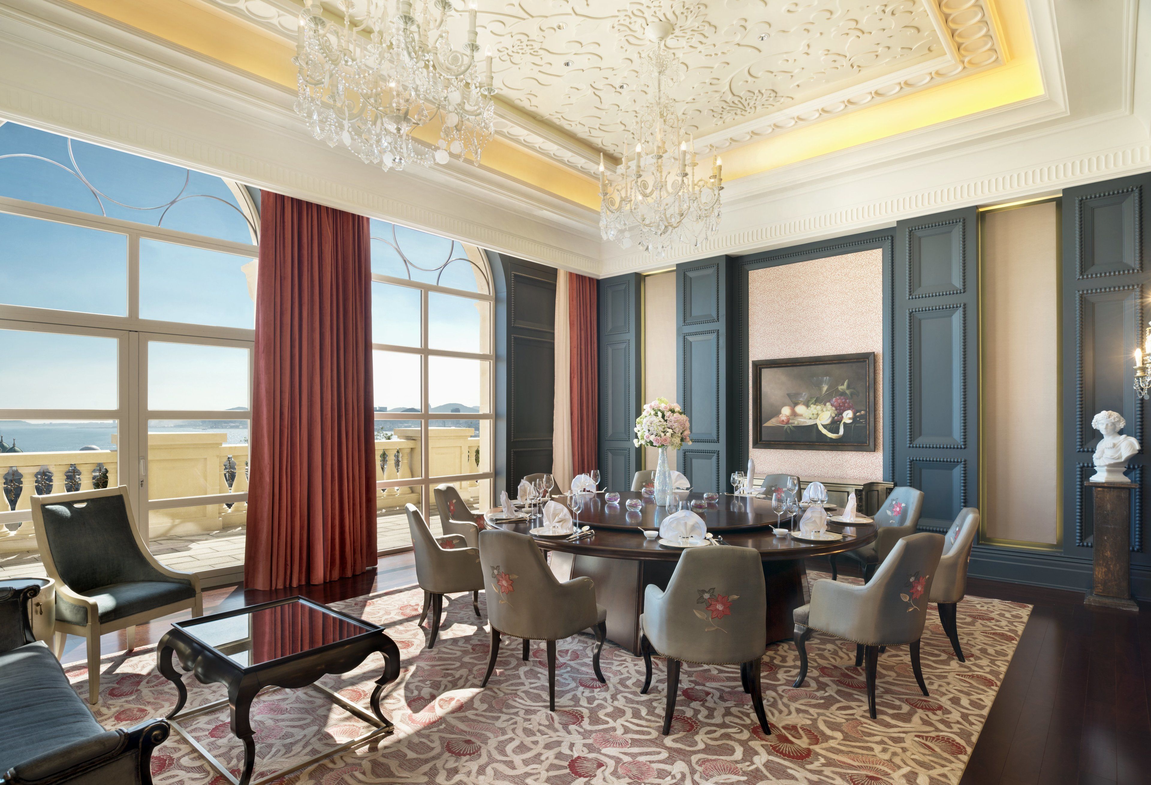 Una habitación lujosa con una gran mesa de comedor preparada para seis personas, rodeada de elegantes sillas. Un candelabro cuelga del techo, y un amplio ventanal ofrece vistas al mar.