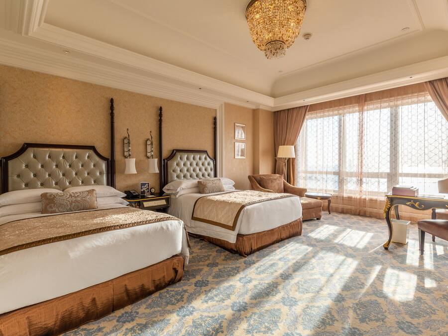 A hotel room with two large beds, a chandelier, a large window with curtains, and a patterned carpet.