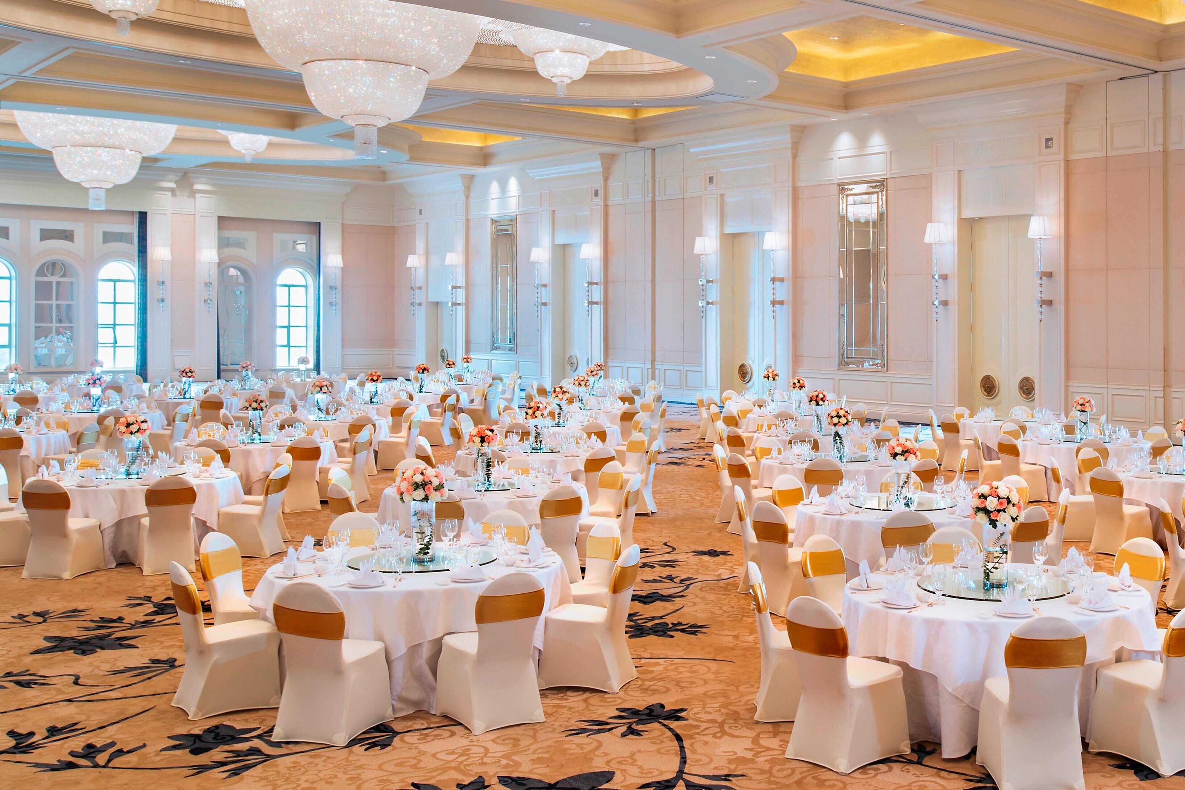 Un gran salón de banquetes con mesas redondas, manteles blancos y fajas doradas.