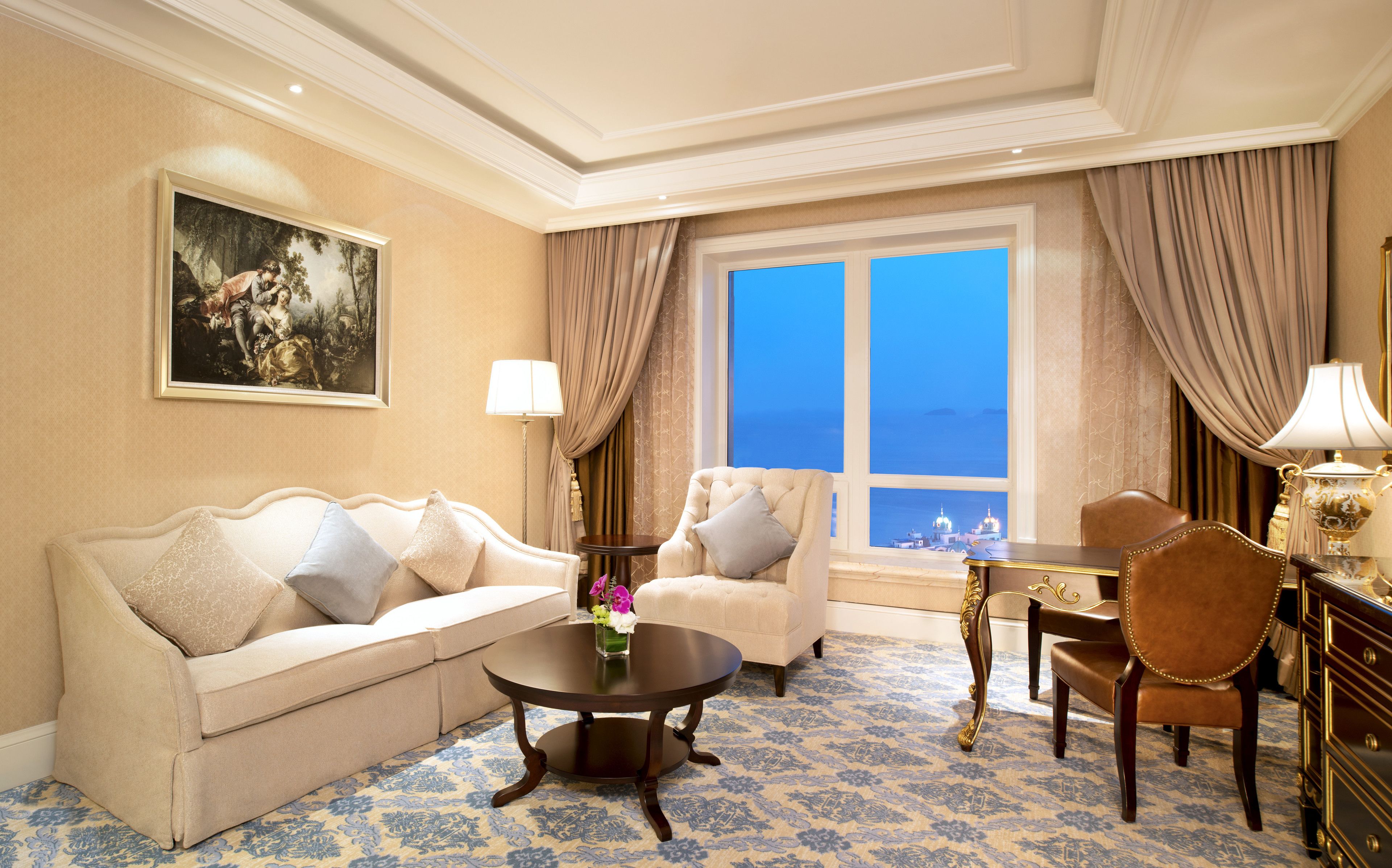 A hotel room with a sofa, armchairs, a dining table, and a view of a cityscape through a large window.