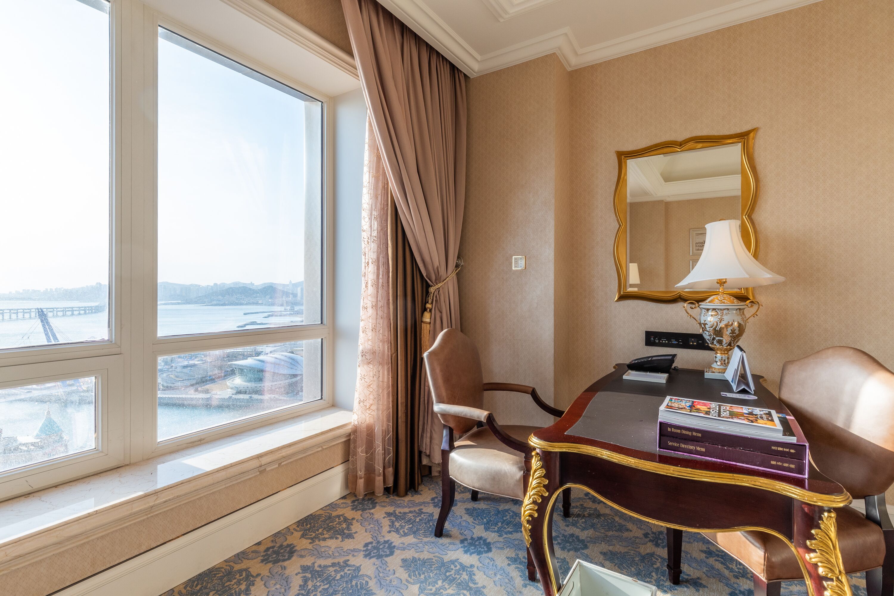 A hotel room with a large window, a desk with a lamp, a chair, and a mirror.