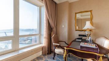 A hotel room with a large window, a desk with a lamp, a chair, and a mirror.