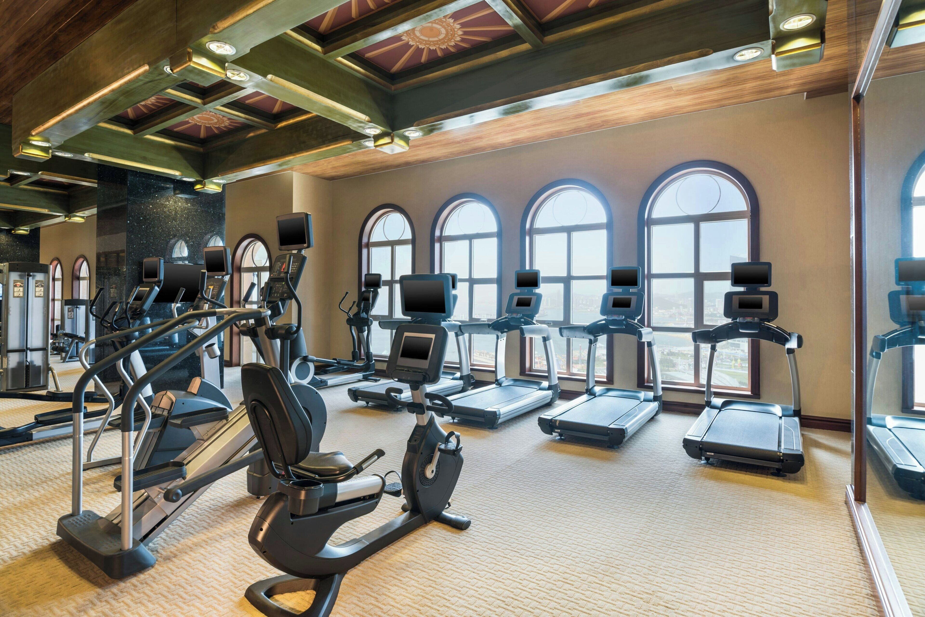 A gym with treadmills, elliptical machines, and stationary bikes, large windows, and a high ceiling with exposed beams.