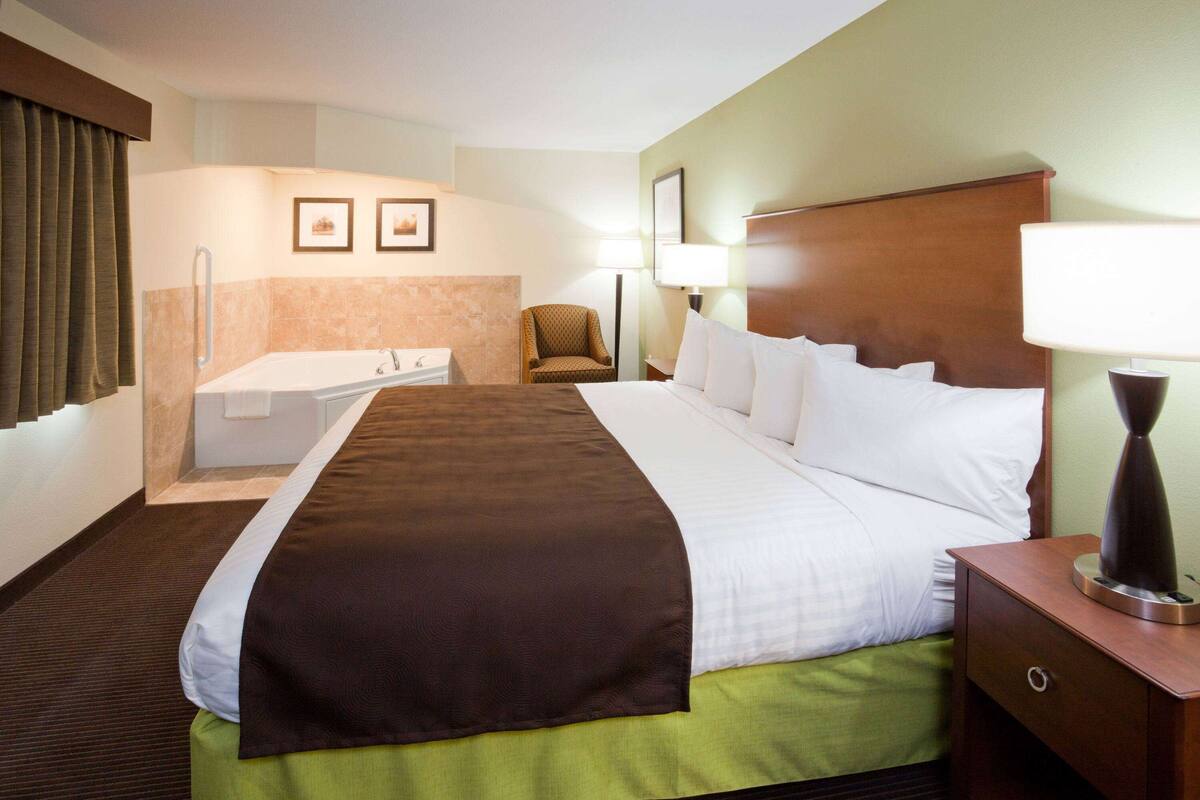 Habitación de hotel con una cama grande, una mesita de noche, una silla, un escritorio, una lámpara y un baño con bañera y toallero.