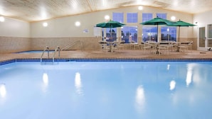 An indoor swimming pool with clear blue water, wooden ceiling, and tiled walls. There are lounge chairs and umbrellas near the pool. - AmericInn by Wyndham Hawley (Hawley)