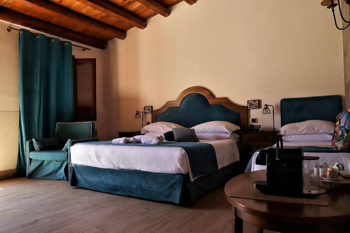 A hotel room with two beds, a wooden ceiling, a green chair, a bedside table with a lamp, and a small round table with a coffee maker.