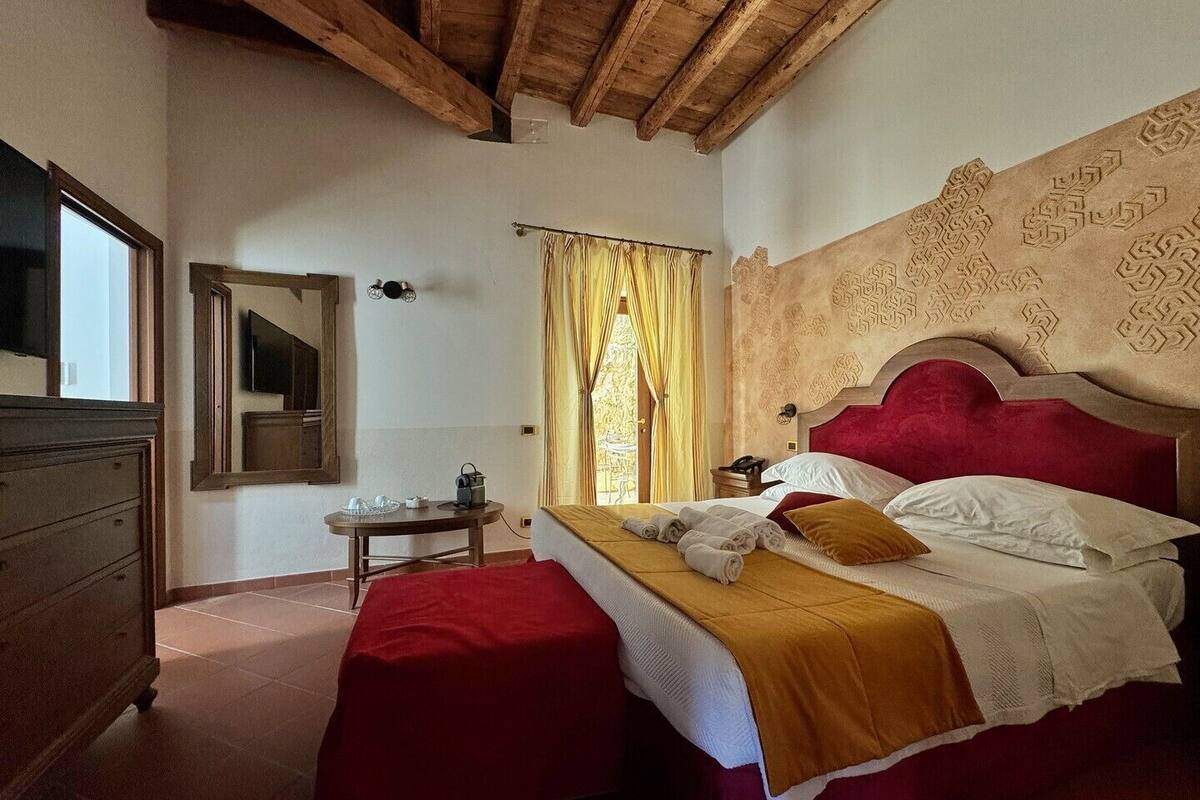 A bedroom with a wooden ceiling, a bed with red and white bedding, a bedside table with a lamp, and a window with curtains.