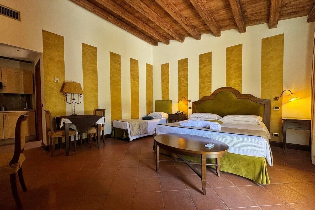 A hotel room with a wooden ceiling, a bed with two pillows, a bedside table, a chair, a small table, and a lamp.