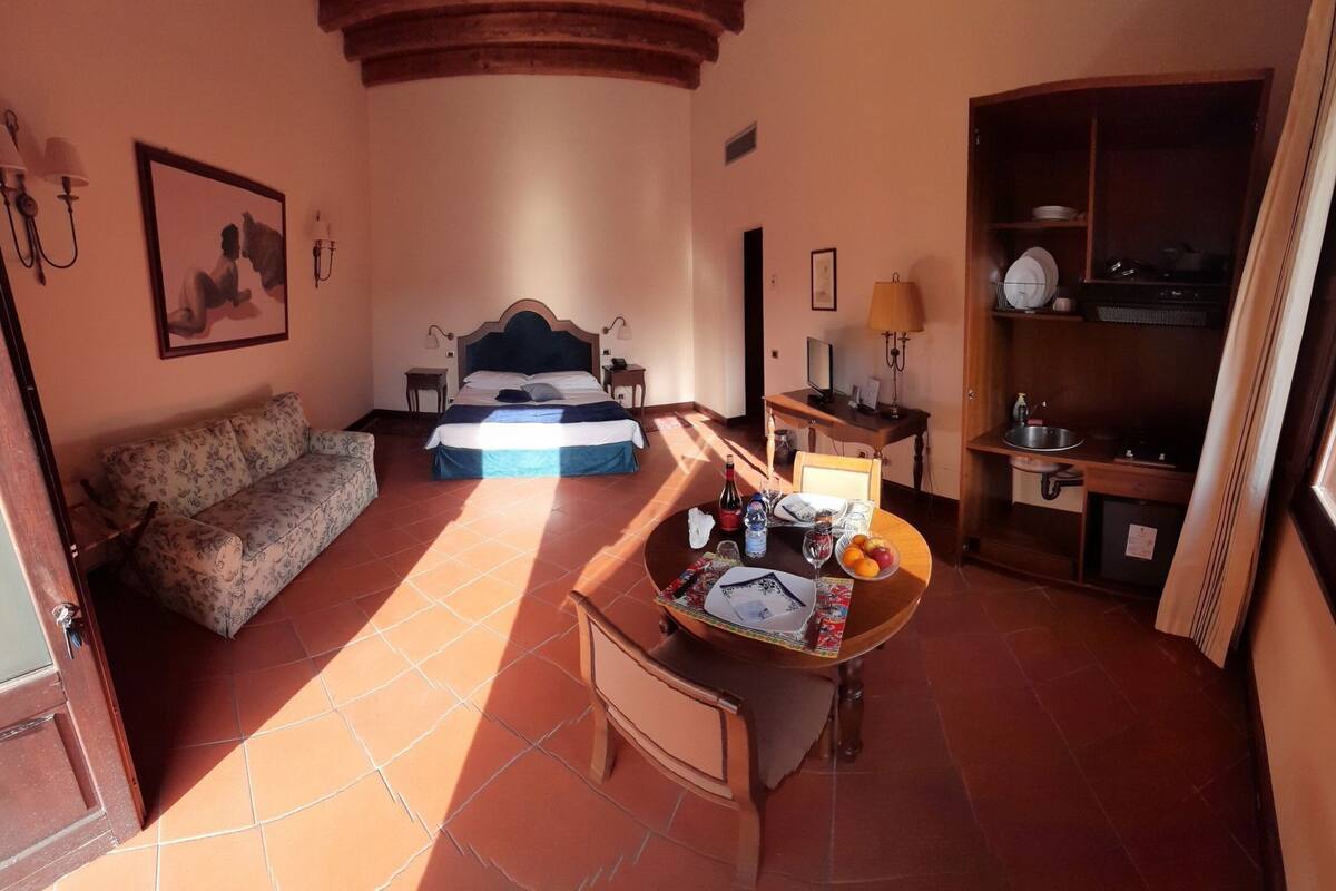 A bedroom with a bed, a sofa, a dining table with a bottle, glasses, and a bowl of fruit.