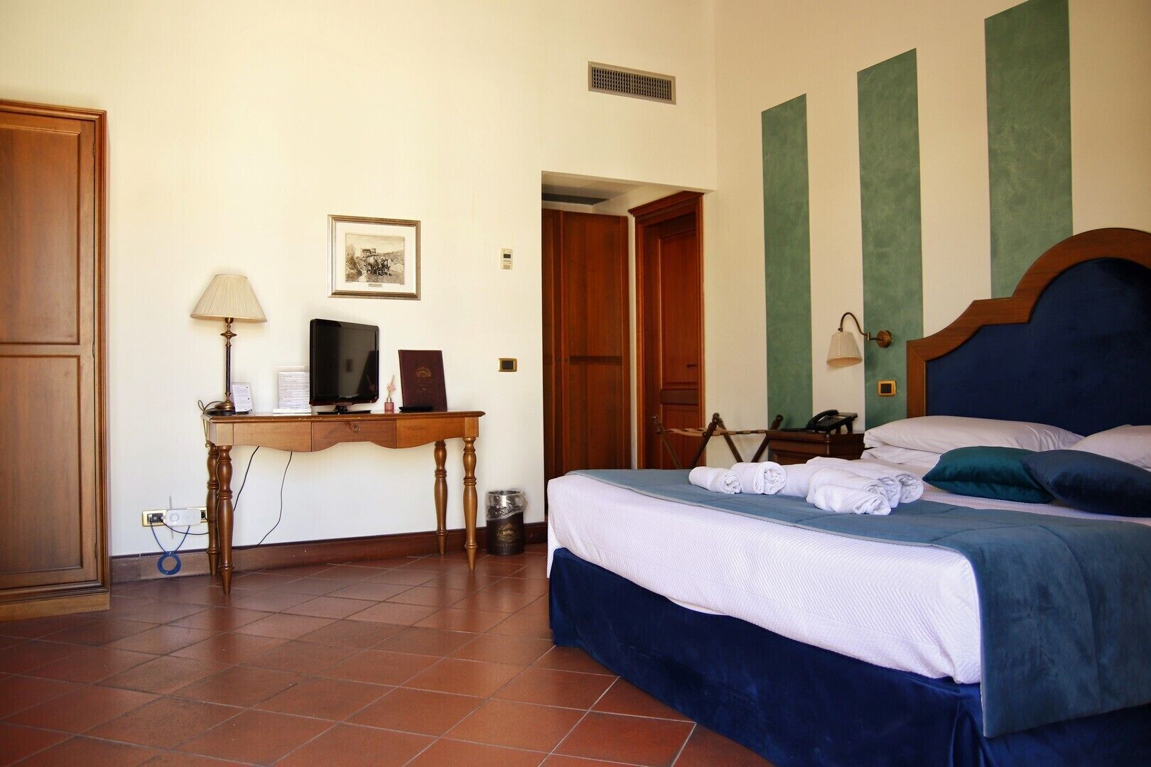 A hotel room with a bed, a desk with a lamp, a television, and a framed picture on the wall.