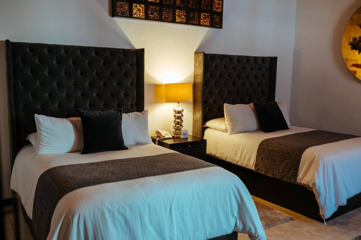 Habitación de hotel con dos camas, cada una con cabecero tapizado, mesita de noche con lámpara y una obra de arte colgada en la pared.