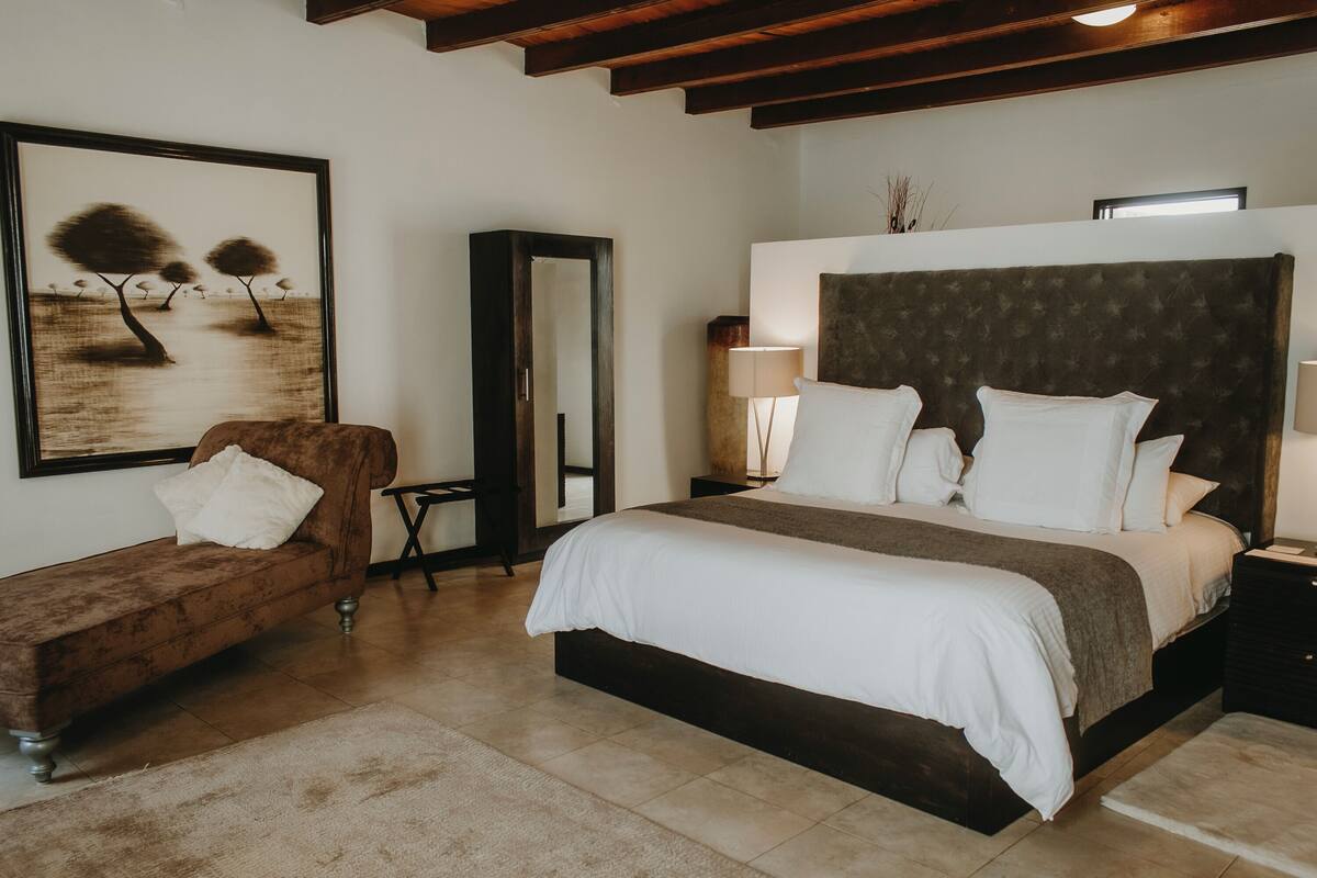 Un dormitorio con una cama grande, un sofá marrón, un espejo y un cuadro con árboles.