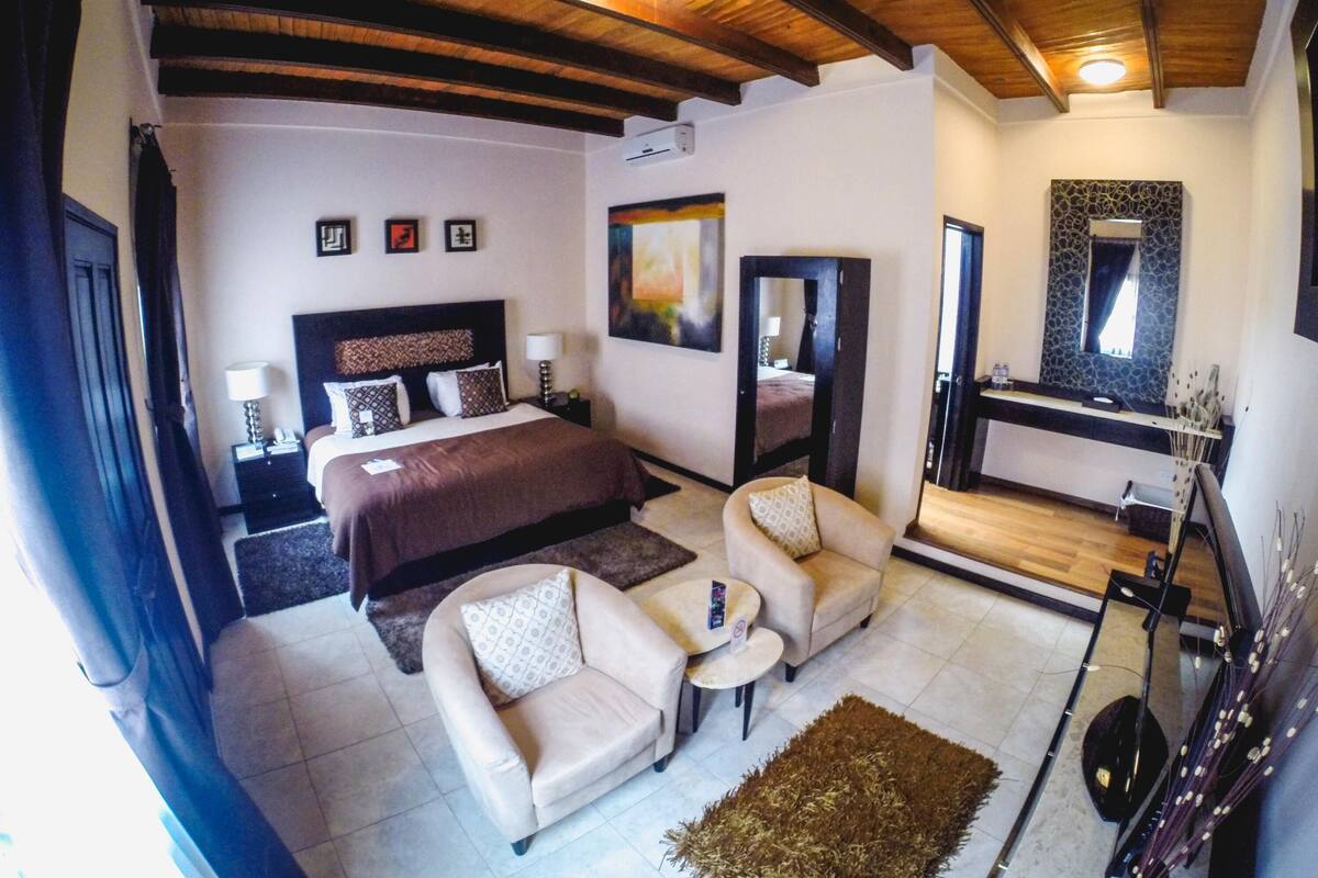 Habitación de hotel con cama, dos sillones, una mesita, un espejo y un baño visible a través de una puerta abierta.