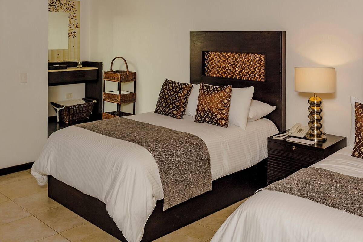 Una habitación de hotel con una cama grande, una mesita de noche con lámpara, una mesita pequeña con una cesta y un estante en la pared.