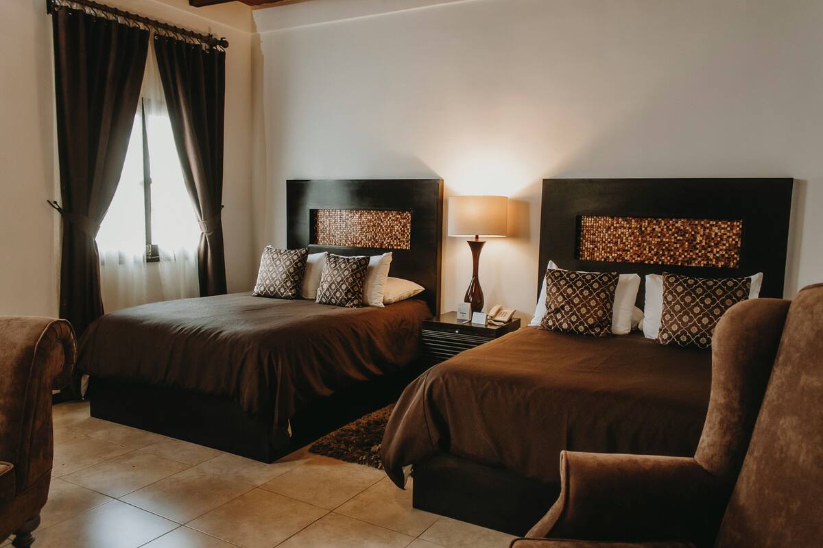 Habitación de hotel con dos camas, una mesita de noche, una lámpara y una ventana con cortinas.