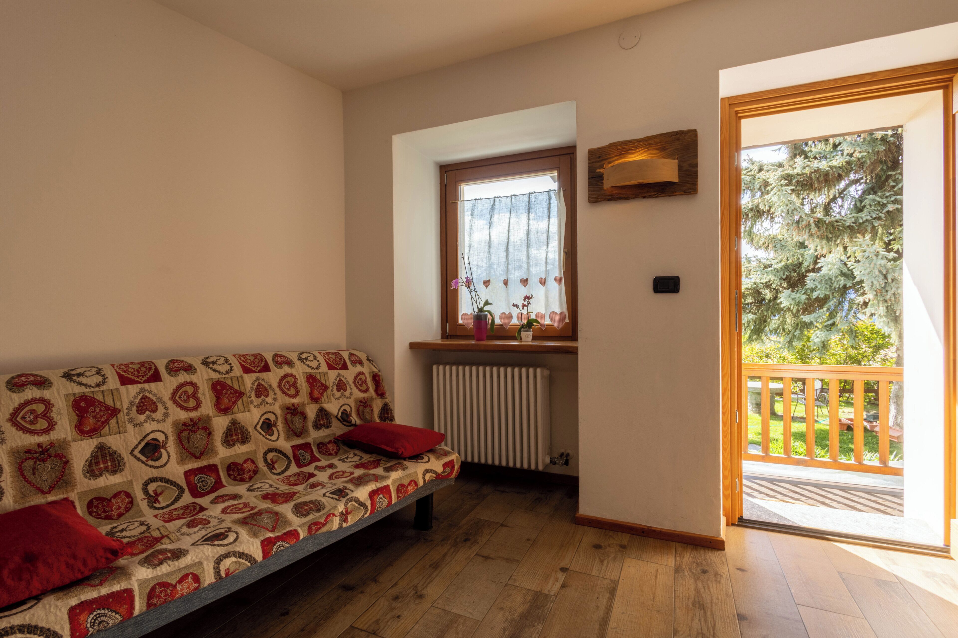 A room with a patterned sofa, a radiator, a window with a curtain, and a view of a wooden deck and trees.
