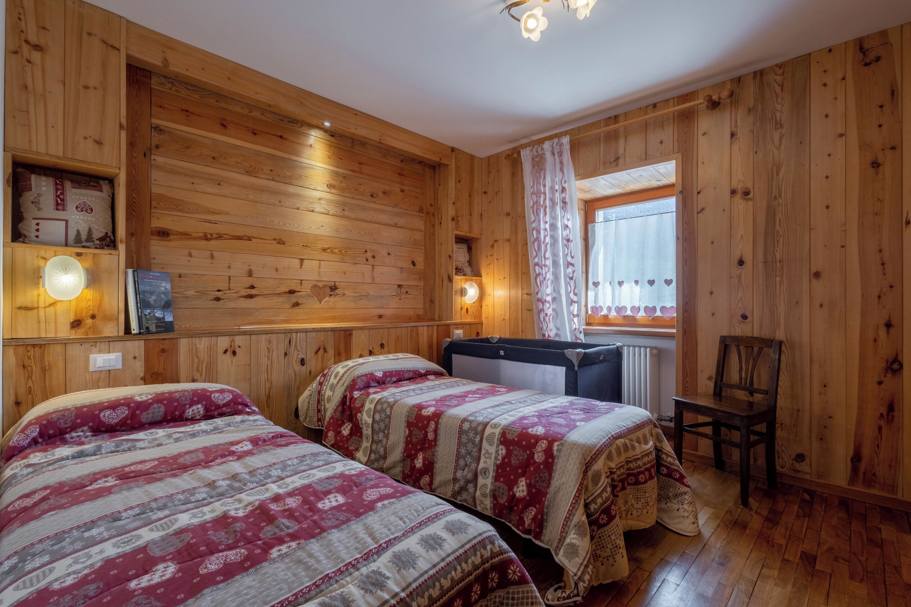 Habitación con paneles de madera, dos camas, una silla, un radiador y una ventana con cortinas de diseño de corazón.