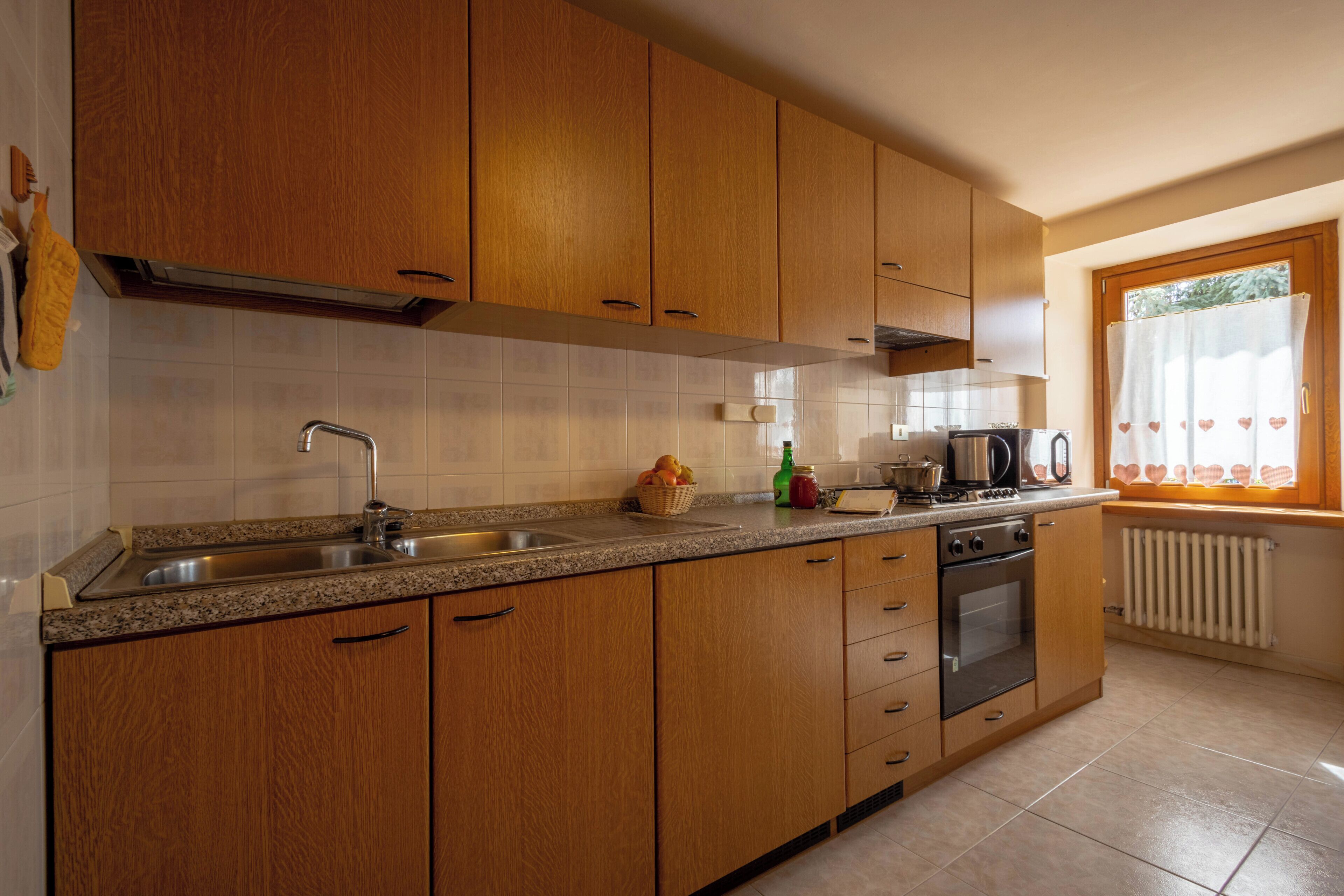Une cuisine avec des meubles de rangement en bois, un évier en acier inoxydable, une cuisinière et une fenêtre avec des rideaux.
