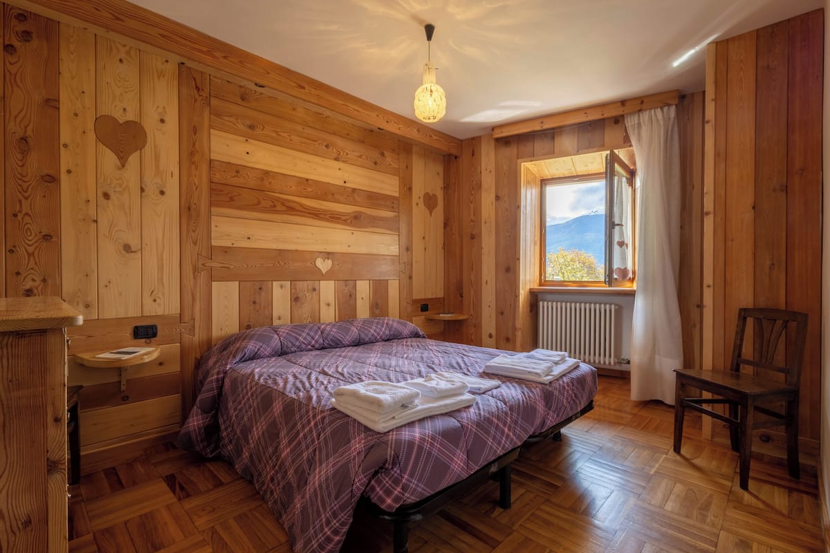 A wooden room with a bed, a chair, a window with curtains, and a radiator.
