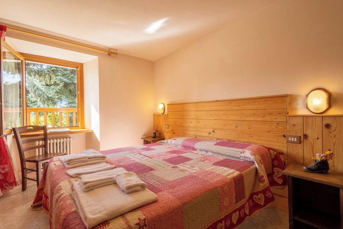 A bedroom with a wooden headboard, a quilted bedspread, a chair, a nightstand with a lamp, and a window with a view of trees.