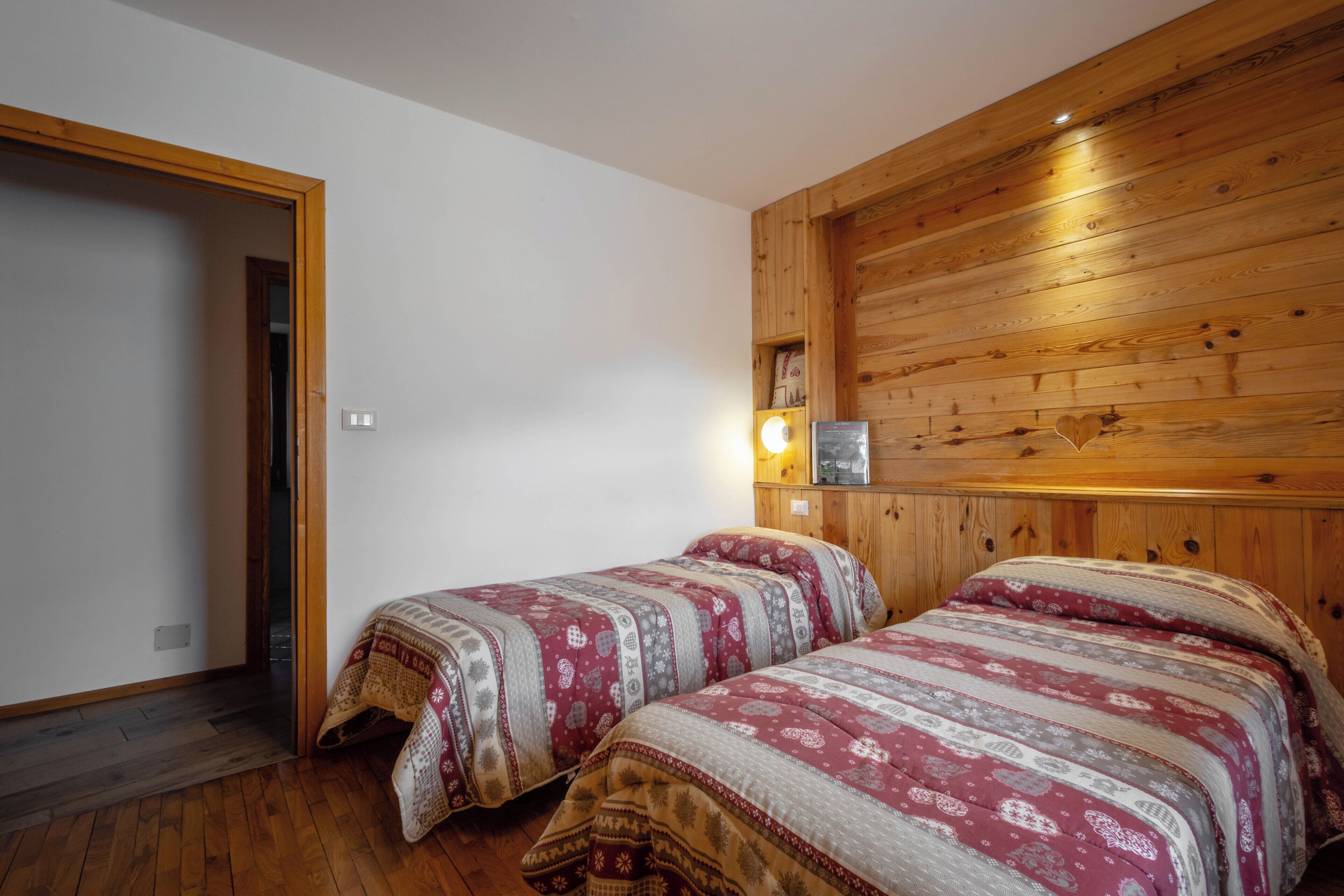 A room with two beds, wooden paneling on the walls, a small shelf with items, and a door leading to another room.