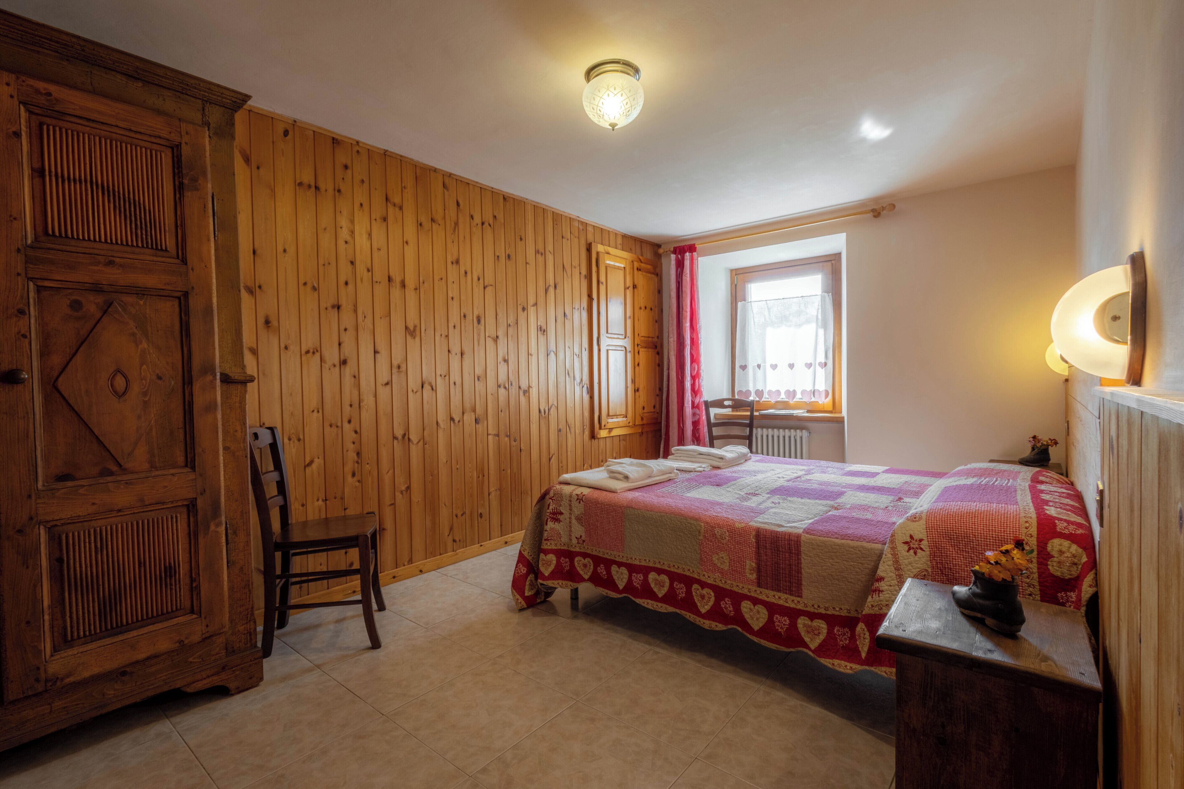 Une pièce aux murs lambrissés, un lit recouvert d’une courtepointe, une chaise, un radiateur et une fenêtre avec des rideaux.