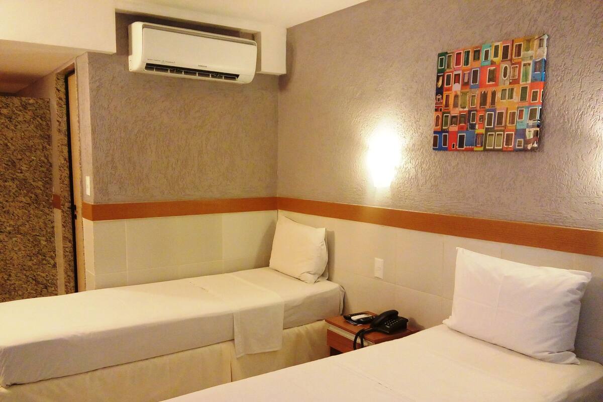 A hotel room with two single beds, a wall-mounted air conditioner, a small bedside table with a phone, and a colorful artwork on the wall.