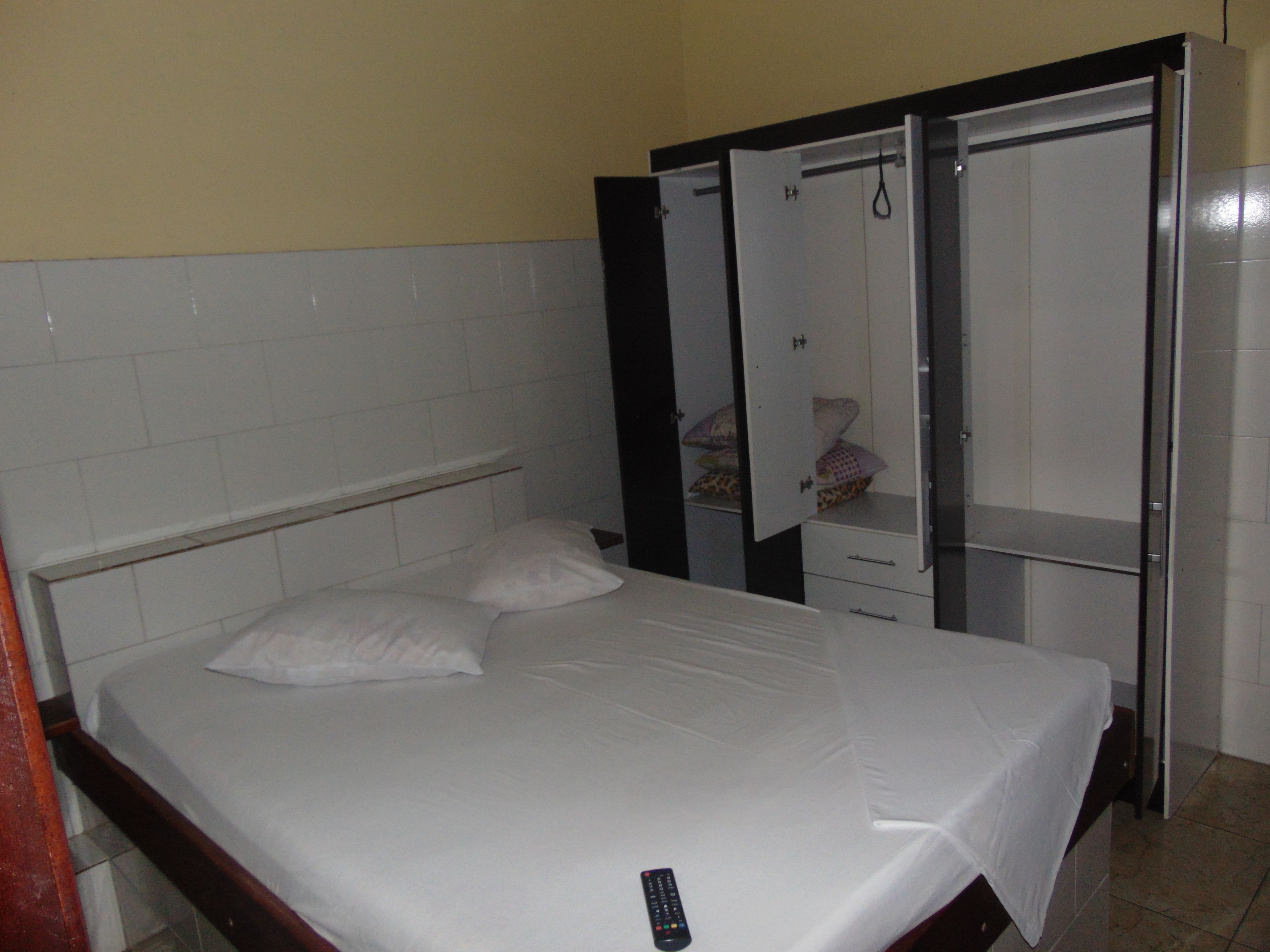 Uma cama de solteiro com colchão e travesseiros brancos, uma mesinha de cabeceira e um guarda-roupa com espelho.