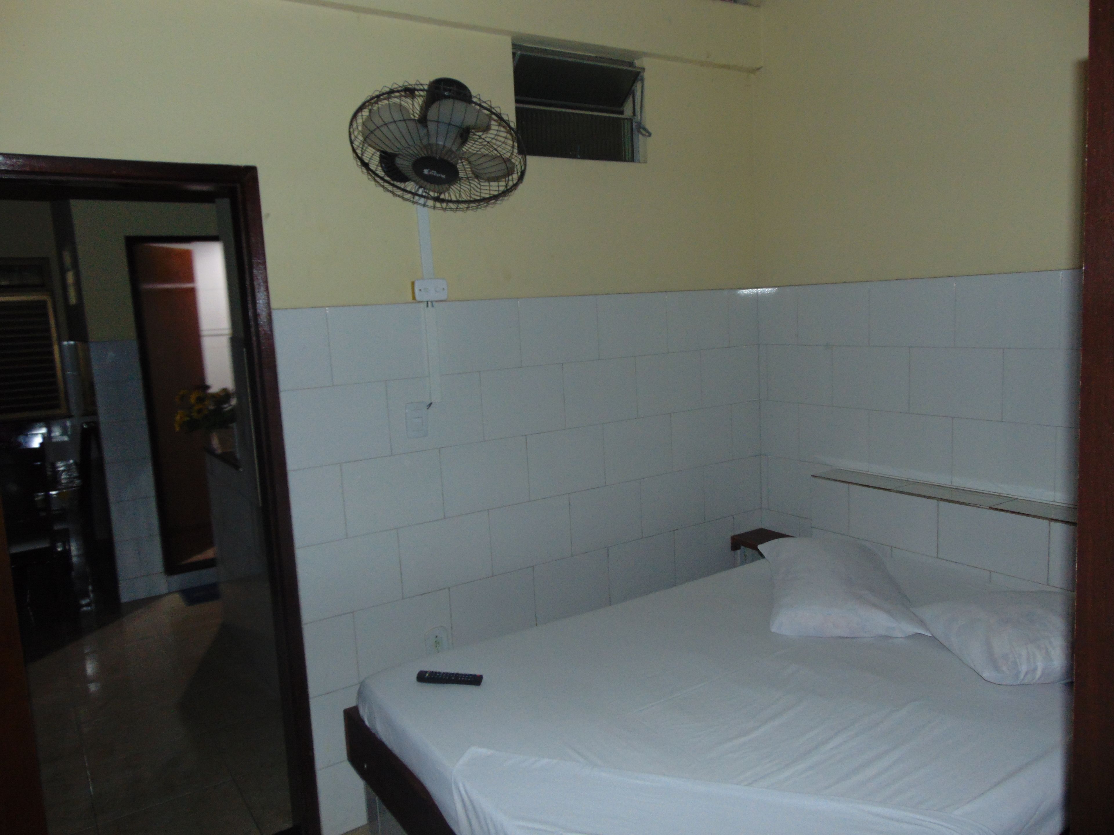 Quarto pequeno com uma cama de solteiro, parede de azulejos brancos, ventilador de teto, janela e porta de acesso a um banheiro.