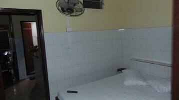 Quarto pequeno com uma cama de solteiro, parede de azulejos brancos, ventilador de teto, janela e porta de acesso a um banheiro.
