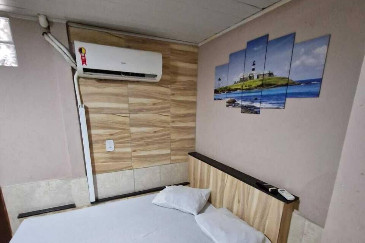 Ein Einzelbett mit Holz Kopfteil und weißer Matratze, eine Wand-Klimaanlage und ein Wandbild mit Leuchtturm und Landschaft.