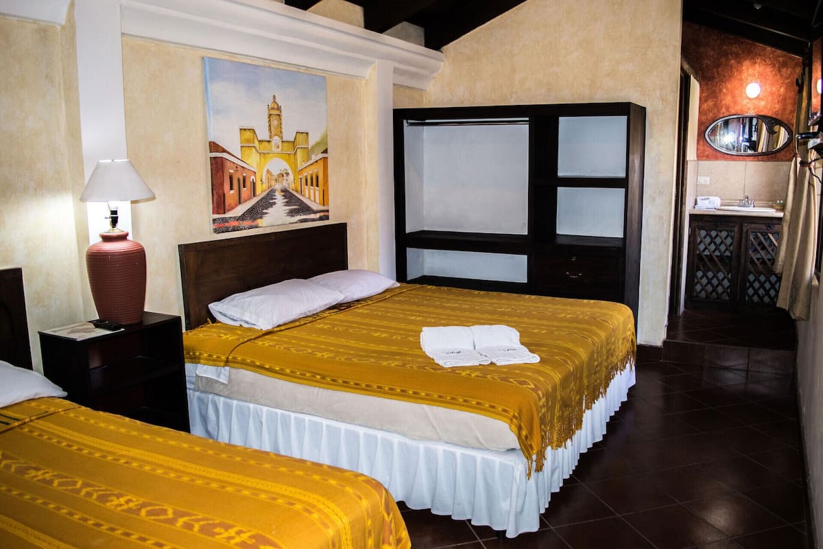 Une chambre d’hôtel avec deux lits, un tableau représentant un bâtiment, un miroir et une salle de bain avec un lavabo et des toilettes.